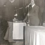 An old photo of a man speaking behind a microphone stand
