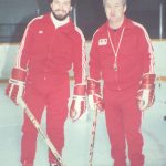Two men holding hockey sticks smiling at the camera on an ice rink