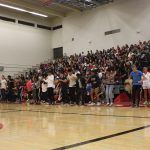 Several students jumping for an activity with other students in the indoor gym seating